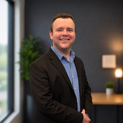 Smiling man in a blue shirt and black blazer standing in a modern office with a window and plants in the background.
