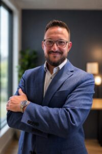 Portrait of a man with glasses and a beard in a blue suit, arms crossed in a modern office setting.