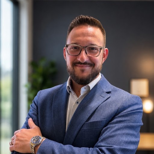 Portrait of a man with short dark hair and a beard, wearing a blue blazer and glasses, smiling with arms crossed at the camera.